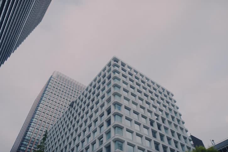 A low-angle, forward-moving shot from a vehicle captures the towering modern skyscrapers and architecture of a city's financial district against an overcast sky