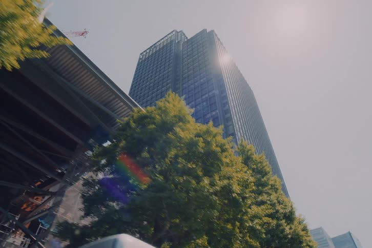 A slow-motion, low-angle shot driving past a modern glass skyscraper and lush green trees on a bright sunny day