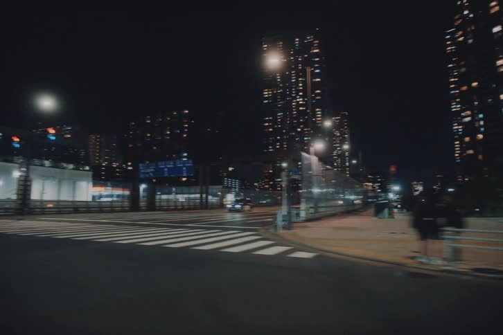 A cinematic tracking shot follows cars driving through a modern city intersection at night, with illuminated skyscrapers creating a vibrant urban backdrop