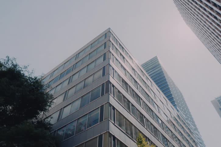 A low-angle point-of-view shot driving through a modern city, looking up at skyscrapers and passing by sunlit trees
