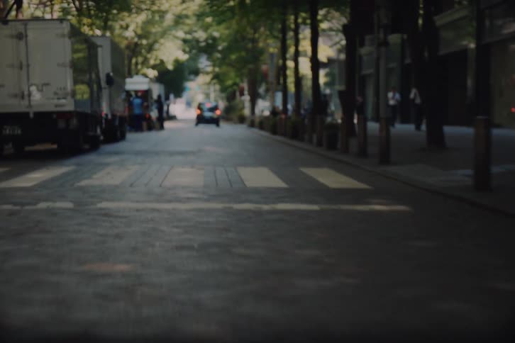 A dynamic point-of-view journey through a modern city street, starting low on the pavement and tilting up to reveal towering skyscrapers and lush green trees