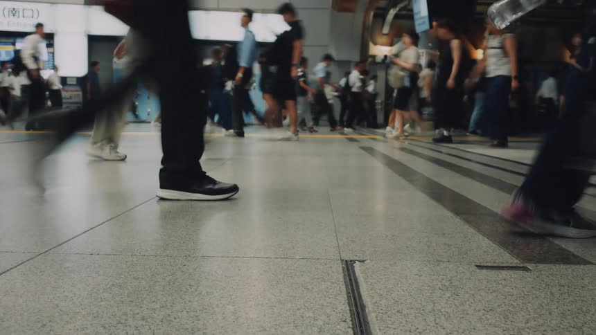 A low-angle time-lapse captures the blurred motion of commuters' feet walking through a busy train station in Japan