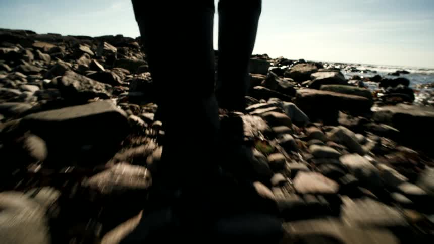 A low-angle, point-of-view shot follows a silhouetted person walking over a rocky beach towards the ocean on a sunny day