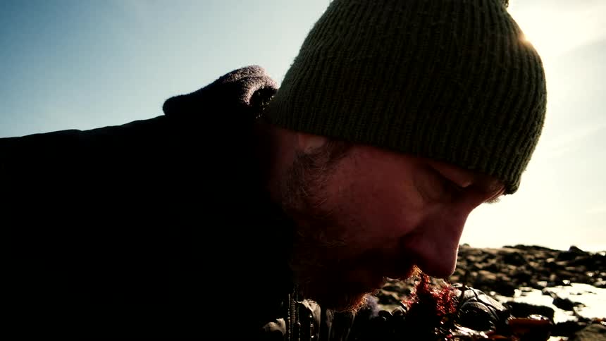 A slow-motion close-up shot shows a bearded man in a beanie smelling freshly foraged seaweed on a rocky beach