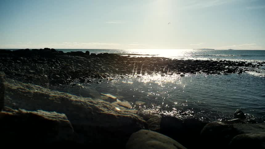 A low-angle view shows the bright sun reflecting on the ocean waves along a tranquil rocky shore
