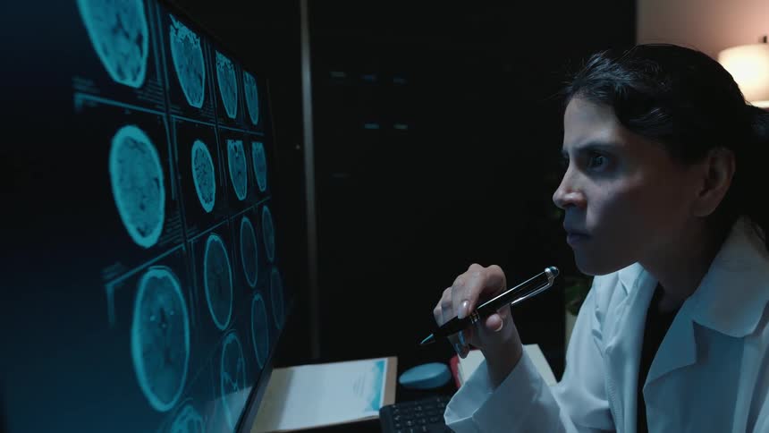 A dedicated female doctor in a lab coat intently studies brain scans on a large monitor in a dark room at night