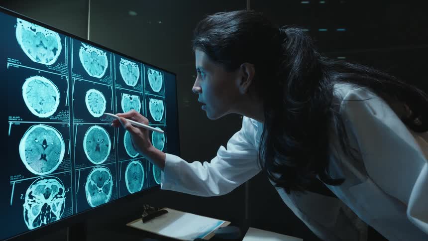 Cinemagraph of a dedicated female doctor intently studying a patient's CT brain scans on a large monitor in a dark, high-tech lab