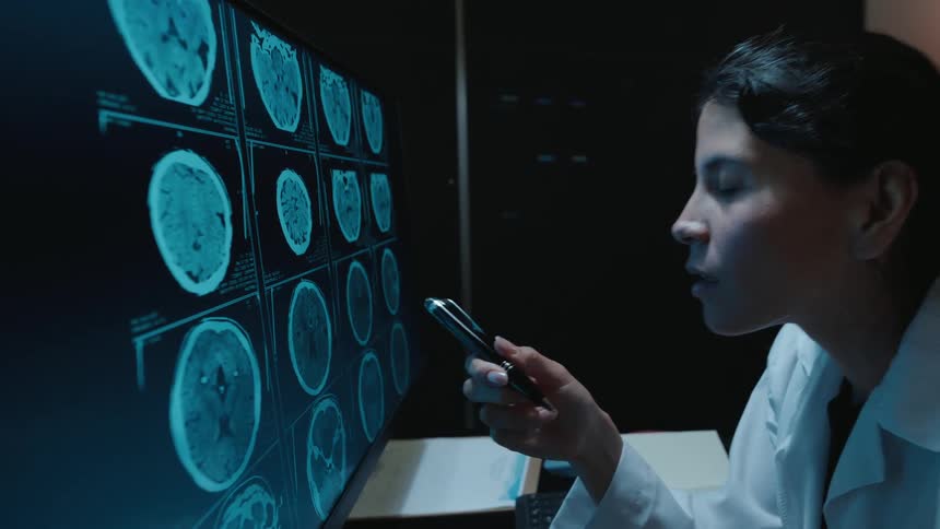 A female radiologist in a dark room intently analyzes a patient's CT brain scans on a monitor before making a discovery and taking notes