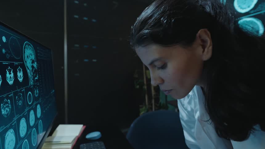 A female doctor in a lab coat analyzes brain scans on a large screen and computer monitor in a dark medical office