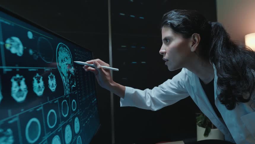 A dedicated female doctor works late, examining a patient's brain MRI scans on a futuristic digital interface