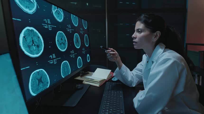 A female doctor in a lab coat meticulously examines brain scan images on a large computer monitor in a dark office