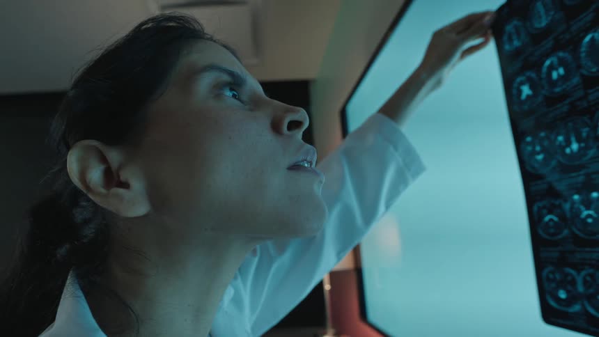 A dedicated female doctor in a dark room meticulously examines a patient's brain MRI scan on a large light screen
