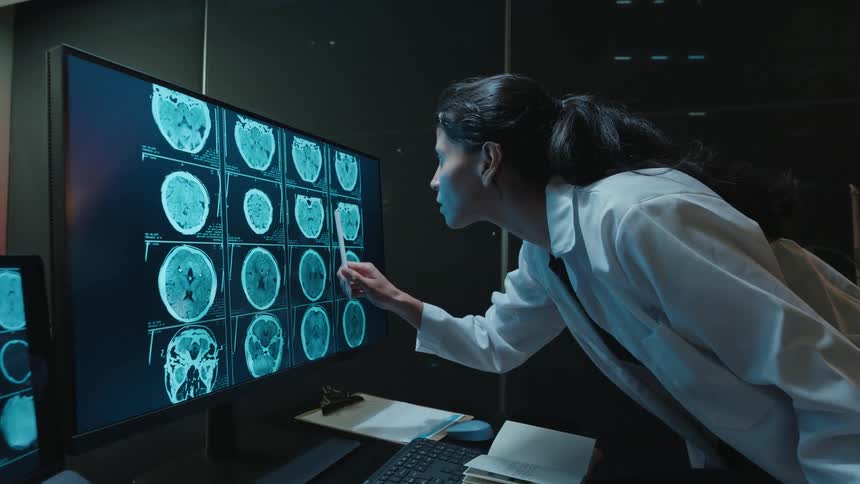 A female doctor meticulously analyzes brain scans on a large computer monitor in a dark, modern research lab