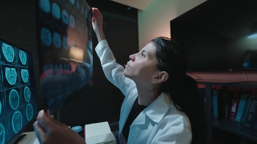 A focused female doctor in a dark office carefully holds a brain scan film up to the light to analyze it