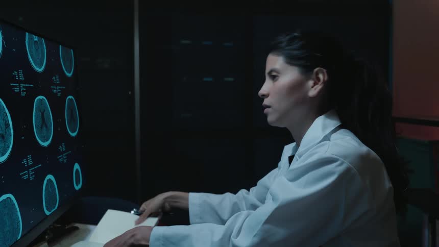 A female doctor in a lab coat carefully analyzes brain scans on a computer monitor in a dark office at night, taking notes on her findings