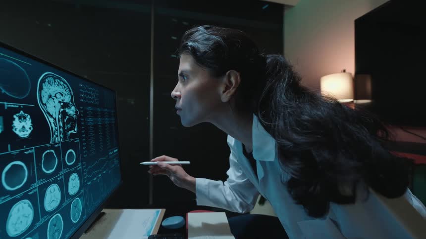 A focused female neurologist in a lab coat closely examines detailed MRI brain scans on a large computer screen in her dark office at night