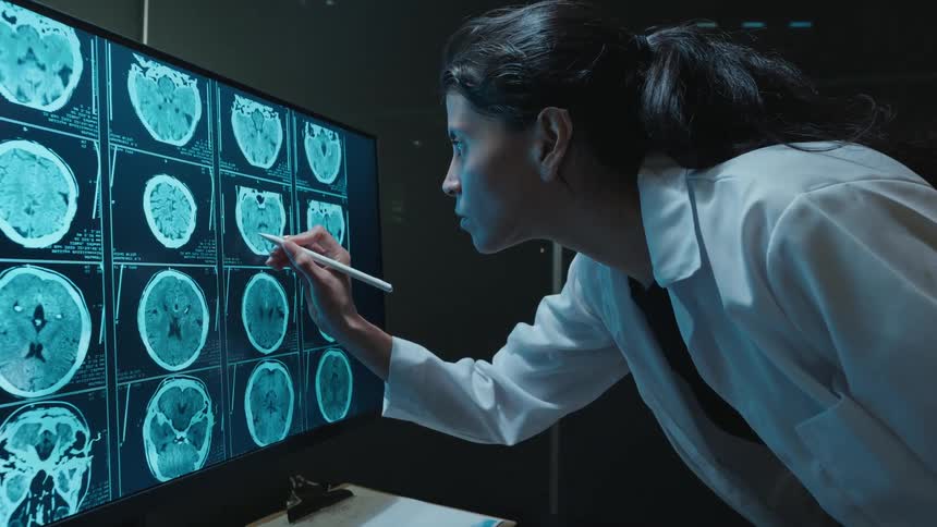 A female medical professional uses a stylus to carefully examine MRI scans of a human brain on a large computer screen