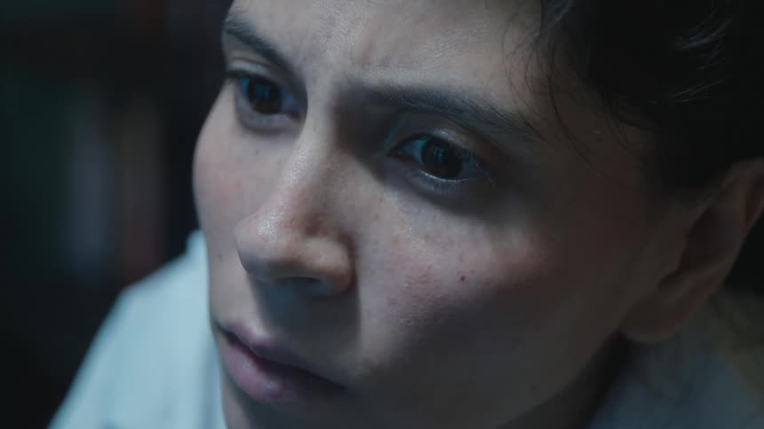 Extreme close-up on a young woman's face as she stares at something off-screen with wide, fearful eyes in a dimly lit room