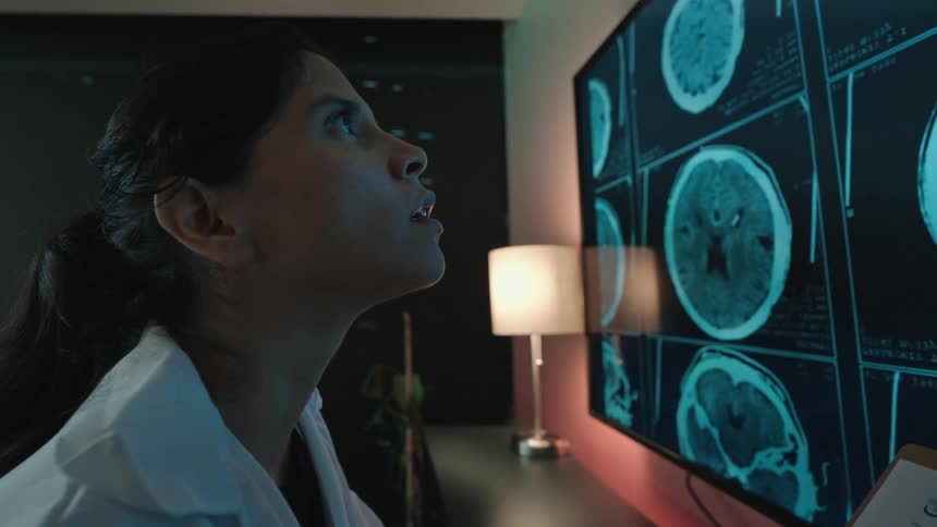 A dedicated female doctor analyzes brain scans on a large screen in a dark office, her focused expression turning to a smile of relief and hope