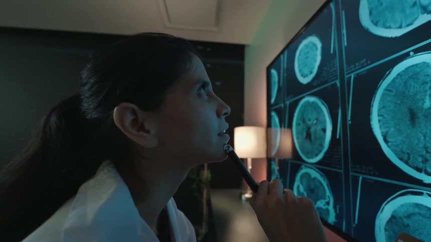Side view of a female medical professional thoughtfully examining brain scans on a high-tech monitor in a dark office