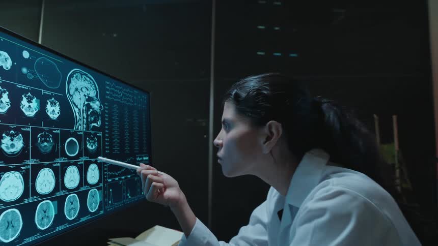 A female scientist in a lab coat studies human brain tomography results on a high-tech computer monitor