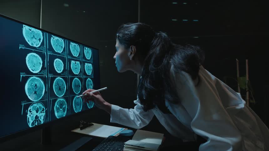 A dedicated female doctor in a lab coat works late at night, meticulously analyzing brain scans on a large computer monitor with a stylus