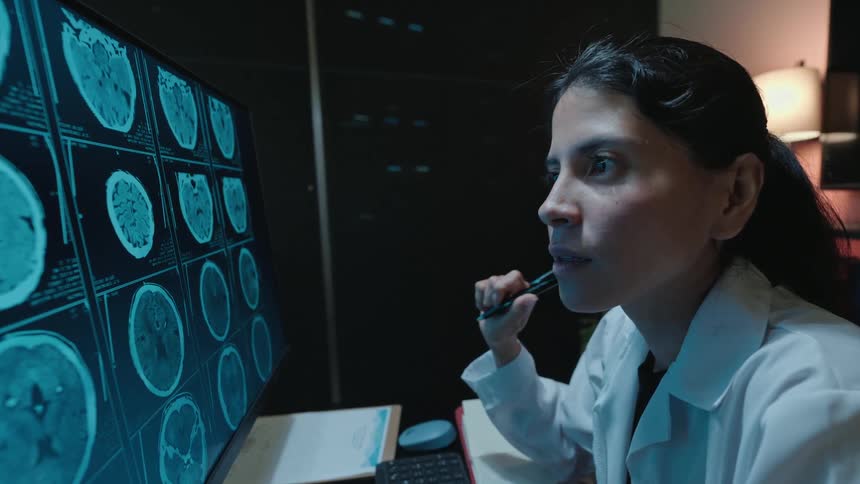 A female doctor in a dark room meticulously studies a series of brain scans on a computer monitor and makes a discovery