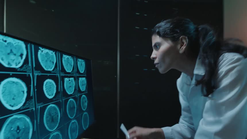A female doctor in a dark room meticulously examines brain MRI scans on a physical film and then on a large computer monitor