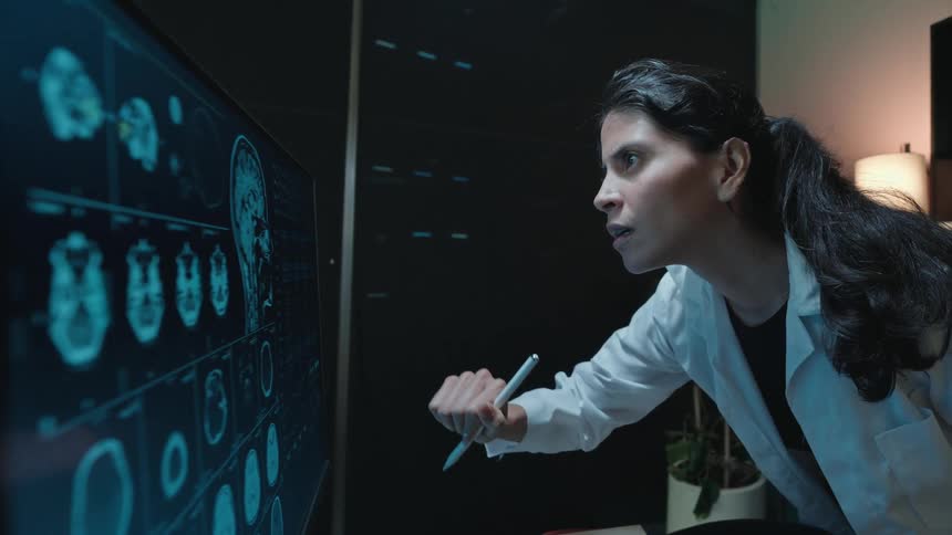 A female medical professional intensely examines brain MRI scans on a large digital screen in a dark laboratory