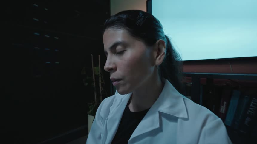 A female doctor in a white coat writes on a clipboard in a dark office and then looks up with a shocked and surprised expression