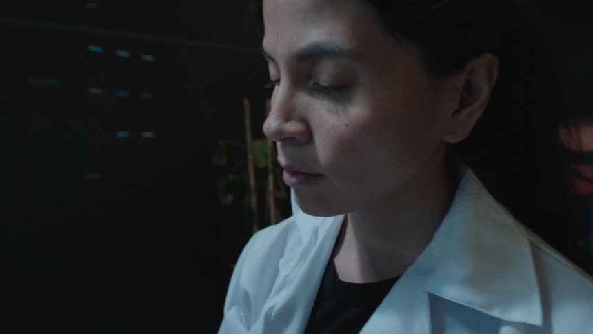 Slow dramatic zoom on a female doctor in a white lab coat writing notes at her desk in a dimly lit office at night