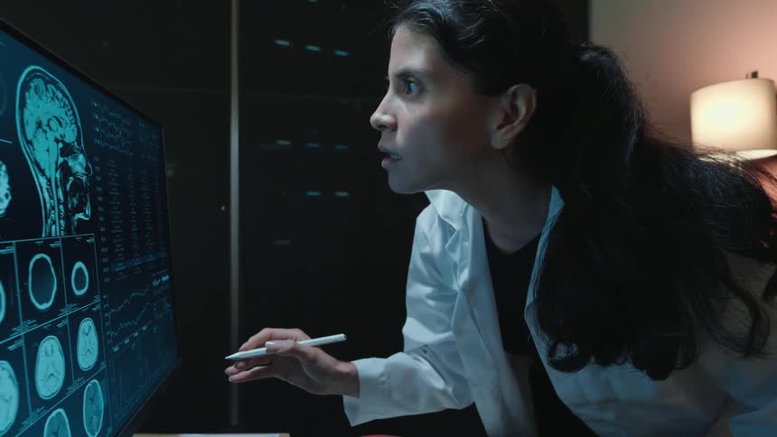 A female doctor in a dark lab intently studies patient brain scans and data on a large, high-tech computer monitor