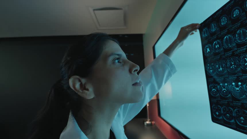 A female doctor in a lab coat closely examines a patient's brain MRI scan on a large light screen in a dark medical office
