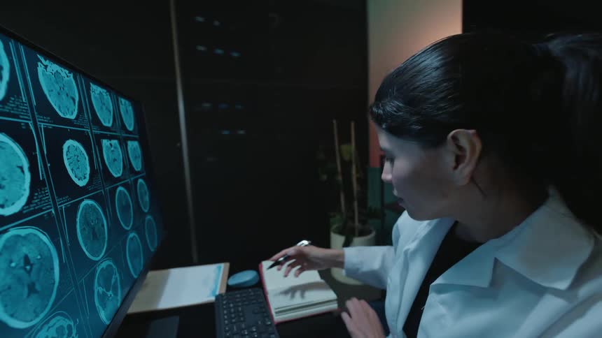 A female doctor in a dark room meticulously examines brain scans on a computer monitor and writes her findings in a notebook