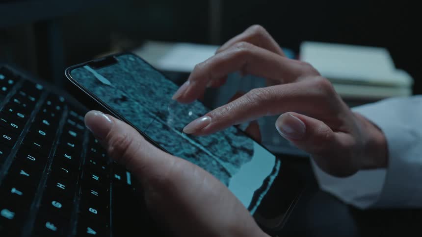 A medical professional meticulously examines a patient's brain CT scan on a smartphone, zooming and scrolling through the layers in a dark room