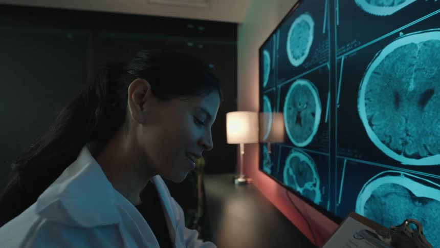 A female neurologist examines a patient's brain scans on a large screen in a dark room, smiling as she makes a diagnosis