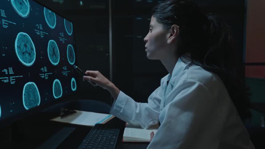 A dedicated female neurologist studies a patient's brain scans on her computer monitor late at night in her modern office