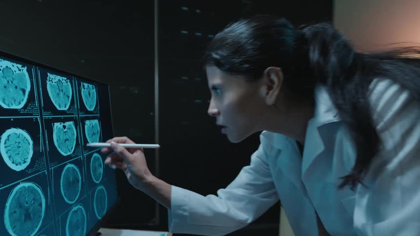 A focused female doctor in a lab coat intensely analyzes brain CT scans on a large computer screen in a dark medical office