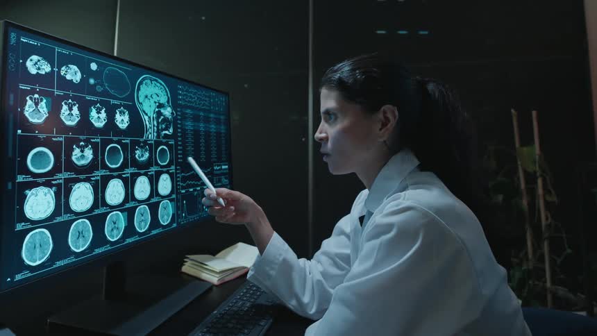 A focused female doctor points with a stylus while meticulously analyzing MRI brain scans on a large computer screen in a modern, dark laboratory