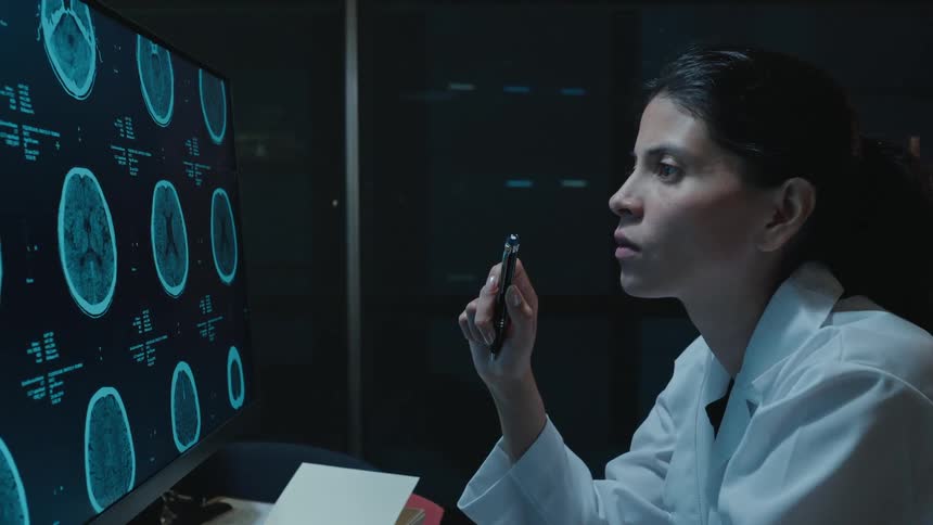 A female doctor in a lab coat studies CT brain scans on a computer monitor while working late in a dark office
