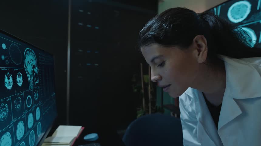 A female neurologist in a lab coat intently studies brain scans on a computer monitor in a dark, high-tech laboratory
