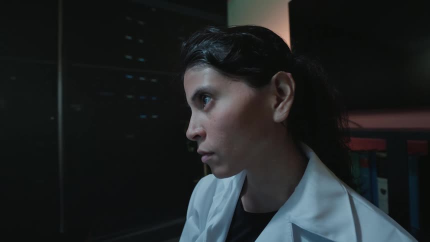 A focused female doctor in a lab coat writes notes while analyzing brain scans on a computer in a dark office