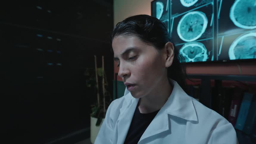 Slow zoom dolly shot of a female doctor in her dark office analyzing patient brain scans with a concerned and focused expression