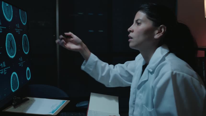 A female neurologist analyzes a patient's brain CT scan on her computer and writes a diagnosis in her notebook