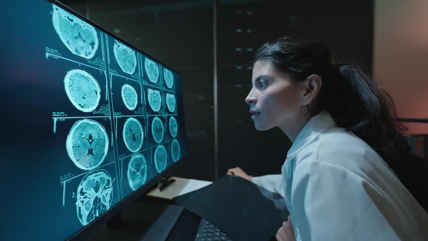 A female doctor in a white lab coat meticulously analyzes brain scans on a film and a computer monitor in a dimly lit office