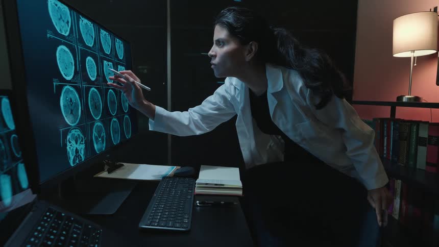 A dedicated female doctor in a white lab coat works late at night in her office, meticulously examining brain scans on a large computer monitor