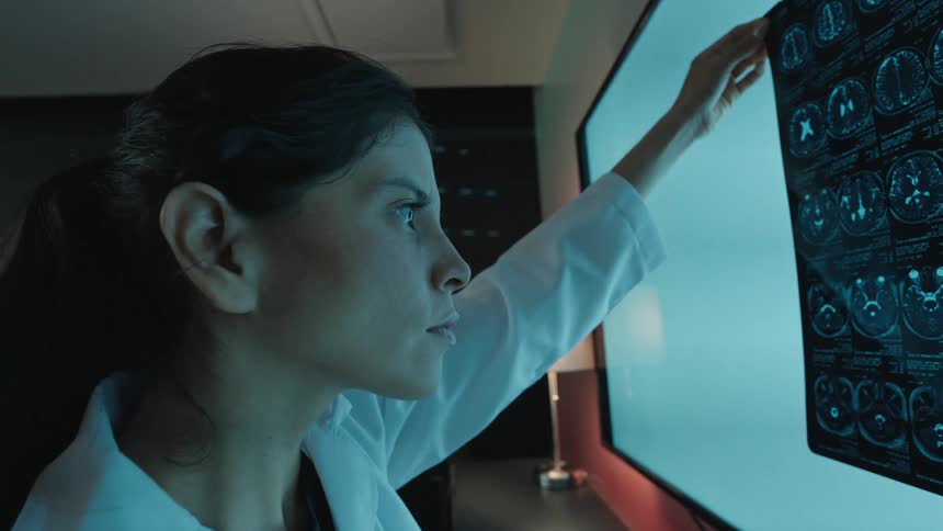 A dedicated female doctor in a dimly lit room meticulously examines a brain MRI scan held against a glowing light panel