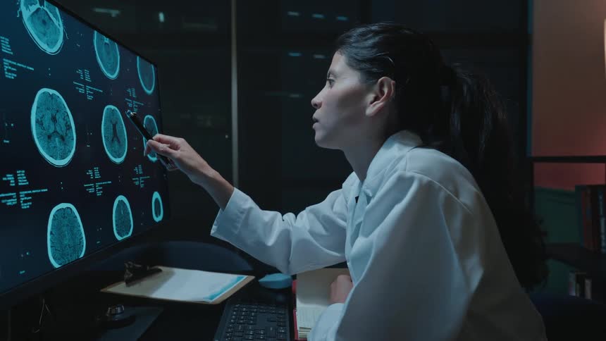 A focused female doctor in a white coat analyzes patient brain scans on a large computer monitor while working late in her office