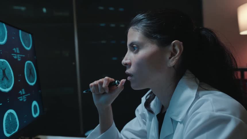 A focused female doctor in a white lab coat thoughtfully analyzes a patient's CT brain scan on a computer monitor in a dark room