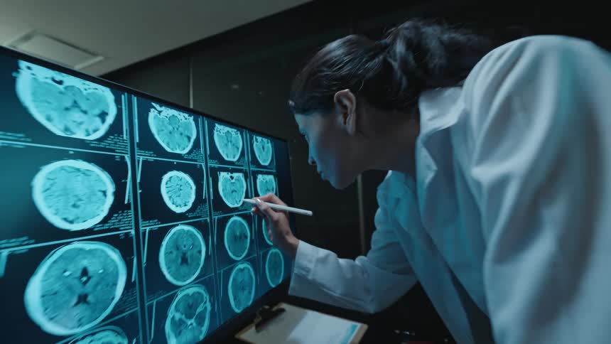 A focused female neurologist points a stylus at a series of brain MRI scans on a large screen in a dark medical office